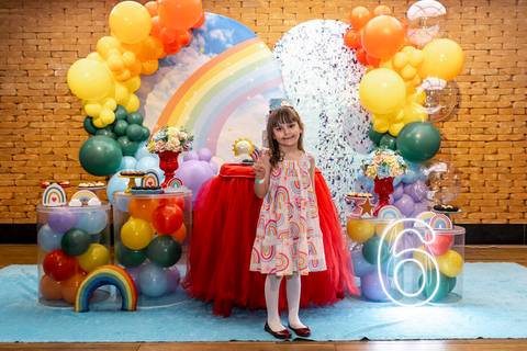 Fotografia de aniversário infantil no Plus Buffet em São Paulo: aniversariante Giovanna sorri em frente à decoração completa da festa com tema arco-íris e o número 6 iluminado.'