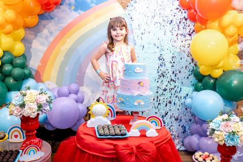Fotógrafo de festa infantil no Plus Buffet registra a aniversariante posando ao lado do bolo de 3 andares, com decoração de balões e painel de arco-íris ao fundo.