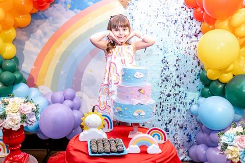 Registro espontâneo em festa infantil no Plus Buffet: menina faz pose divertida atrás da mesa do bolo, em sua festa de 6 anos com tema arco-íris, fotografada em São Paulo.