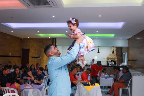 Pai erguendo a filha com alegria durante festa infantil em Guarulhos, fotografia espontânea de família.'