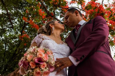 Fotografia dos noivos se olhando, com o buquê em primeiro plano, visto de baixo, com flores de flamboyant no alto da imagem'