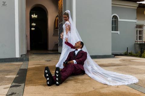 Fotografia da noiva puxando o noivo pela gravata para dentro da igreja, sapato do noivo escrito 