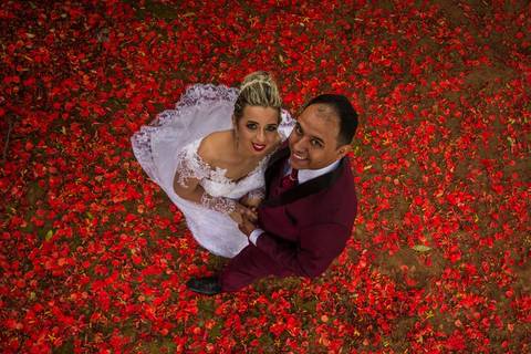 Fotografia dos noivos, vistos de cima, com chão forrado de flores vermelhas durante ensaio fotográfico pós casamento realizado na cidade de Araçuaí, Minas Gerais'