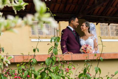 Fotografia dos noivos se olhando, próximo a um arbusto, a noiva segura o buquê e sorri.'