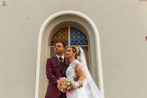 Noivos abraçados, olhando para o horizonte, enquadrados em moldura da porta da igreja'