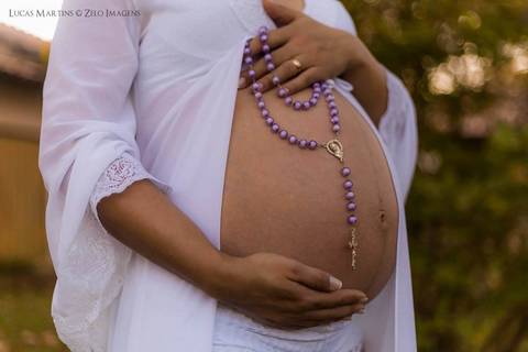barriga da gestante com um modelito branco e um terço roxo'