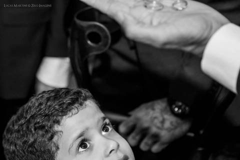 Fotografia de criança, filho da noiva, olhando a palma da mão do celebrante do casamento com as alianças dos noivos.'