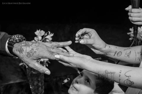 Fotografia de criança fazendo careta olhando enquanto a noiva coloca a aliança no dedo do noivo, ambos tatuados, o noivo usando um relógio, durante casamento realizado no campo no município de Coronel Murta, Minas Gerais.'