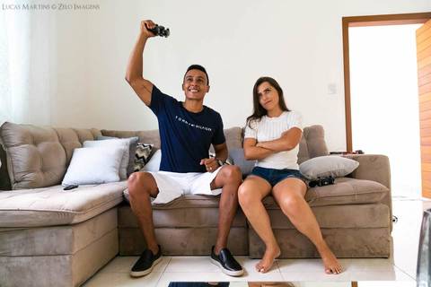 fotografia do casal sentado no sofá, o marido comemorando com o controle de videogame na mão e a esposa brava, durante ensaio fotográfico pós casamento realizado na cidade de Virgem da Lapa, Minas Gerais.'