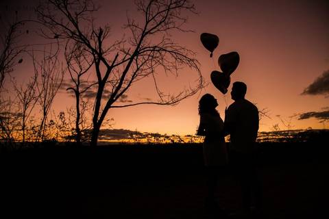 fotografia de silhueta de casal com balões de coração, céu com tom rosado, no fim da tarde em ensaio fotográfico de casal em Araçuaí, Minas Gerais'