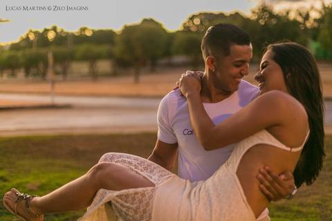 Fotografia de casal. Namorado segura a namorada no colo, os dois se olham sorrindo, durante por do sol, em sessão fotográfica realizada na cidade de Araçuaí - MG'