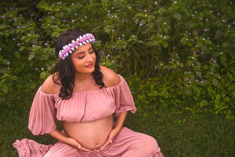 Gestante posando em um gramado verde com um vestido rosa e uma coroa de flores ao fundo com um campo de pequena flores azuis'