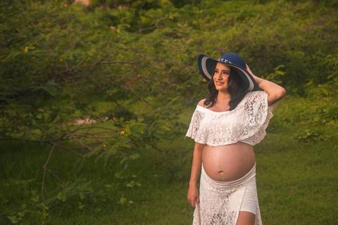 Gestante posando em um gramado verde com um vestido branco e um chapéu azul'