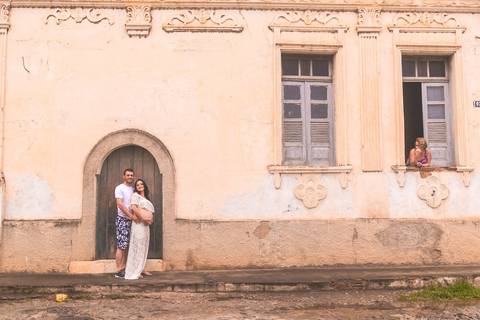 Casal posando em frente uma casa antiga com a dona da casa a janela'