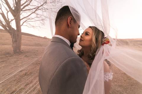 Casal vestindo terno e vestido de casamento enrolados em véu da noiva.'
