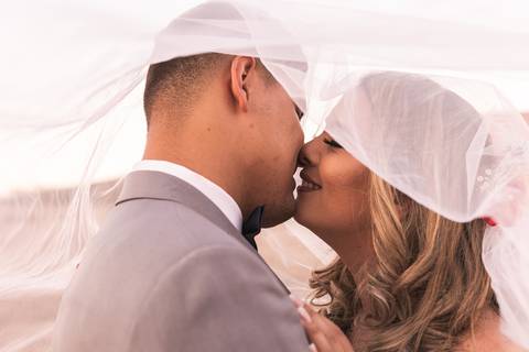 Casal vestindo terno e vestido de casamento enrolados em véu da noiva se beijando. '