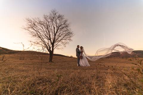 Casal vestindo terno e vestido de casamento posando em campo aberto próximos à uma árvore.'