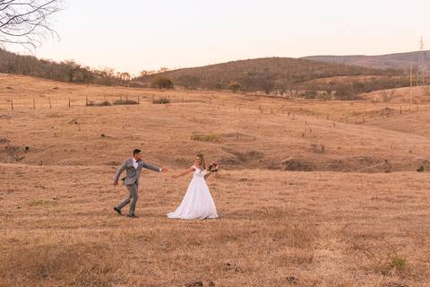 Casal vestindo terno e vestido de casamento correndo em campo aberto.'