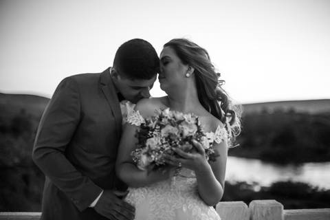Casal vestindo terno e vestido de casamento, o noivo posa beijando suavemente o ombro da noiva.'