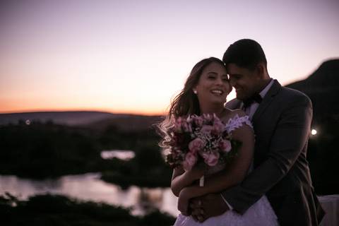 Casal vestindo terno e vestido de casamento posando ao por do sol.'