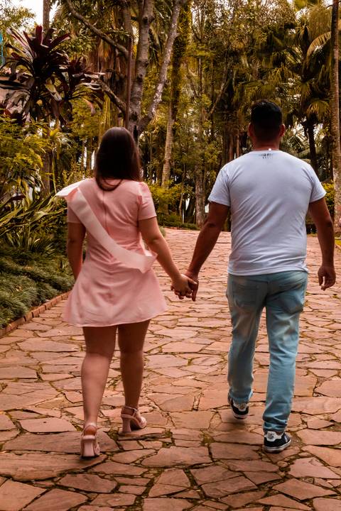 Ensaio pré-wedding, de um casal. Na foto temos um casal, sendo um homem e uma mulher, onde os dois estão de costas, caminhando. A moça está usando um vestido na cor rosa, quase salmão com uma faixa de miss, o homem está de causa jeans azul claro e camisa.'