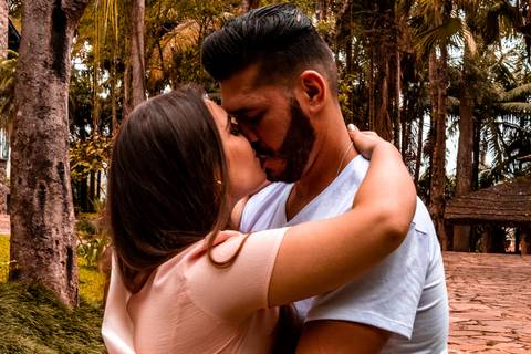 Ensaio pré-wedding, de um casal. Na foto temos um casal, sendo um homem e uma mulher, onde os dois estão se beijando, em um parque. O rapaz está de camisa branca e a moça com um vestido rosa, quase salmão. O parque é uma área verde, com muitas arvores.'