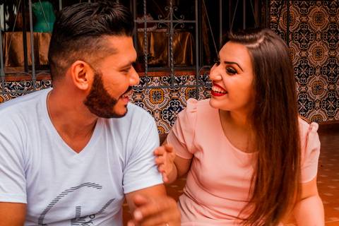 Ensaio pré-wedding, de um casal. Na foto temos um casal, sendo um homem e uma mulher, onde os dois estão sentados um de frente pro outro conversando. A moça está com vestido rosa salmão e o rapaz é barbudo e está com uma camisa branca.'