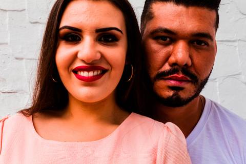 Foto de casal, com enquadramento mais fechado, focado nos rostos do casal, eles estão à frente de uma parede branca. A moça está vestido rosa salmão e o rapaz com uma camisa branca.'