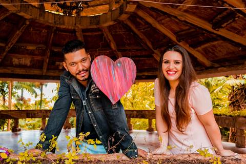 Ensaio pré wedding de casal. os dois estão em sacada de madeira, um ao lado do outro com um coração no meio. o rapaz está de jaqueta e a moça com vestido rosa salmão.'
