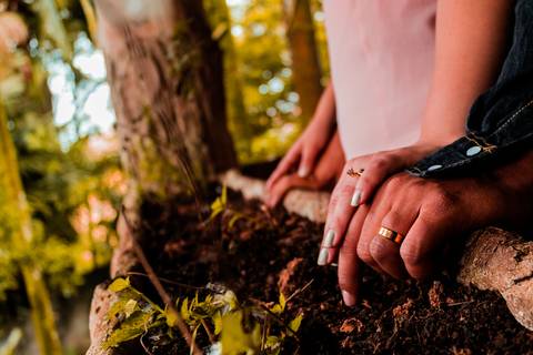 Fotos das mãos do casal em um ensaio pré wedding. As mãos estão sobre uma jardineira de madeira, com uma arvore ao fundo e pequenas flores verdes.'