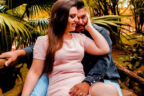 Casal sentado em banco de madeira,
o rapaz está com as pernas abertas e moça está sentada entre elas, com a mão direita no rosto do rapaz. Ela está vestida com vestido rosa salmão e o rapaz com uma jaqueta e causa jeans.'