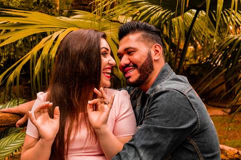 Foto de casal sentados a frente de plantas. Os dois estão descontraídos e sorrindo. Ela está vestida de rosa salmão e ele de jeans.'