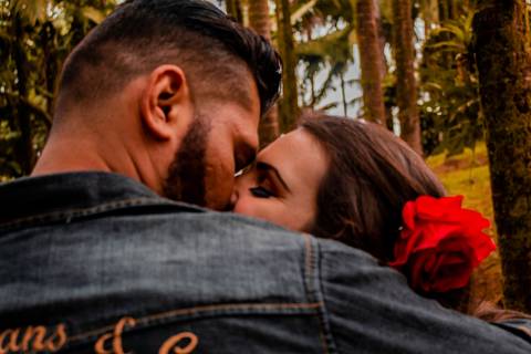 Enquadramento fechado no rosto do casal, que está se beijando. A moça está com uma flor no cabelo e não é possível ver sua roupa, enquanto o rapaz este uma jaqueta jeans.'