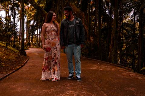 Casal caminhando de frente, em um parque com muitas arvores. a moça veste um vestido longo e florido, enquanto o rapaz usa jaqueta e causa jeans.'