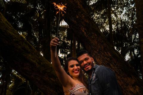 casal em enquadramento do peito pra cima, com as mãos erguidas com um sinalizador acesso. Eles estão á frente de uma arvore. A Moça está vestida com um vestido florido e o rapaz está com jaqueta jeans.'