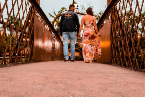 Casal caminhando sobre uma passarela, modelo antigo e na cor marrom. A moça veste um vestido claro e florido, enquanto o rapaz usa uma jaqueta e causa jeans azul.'