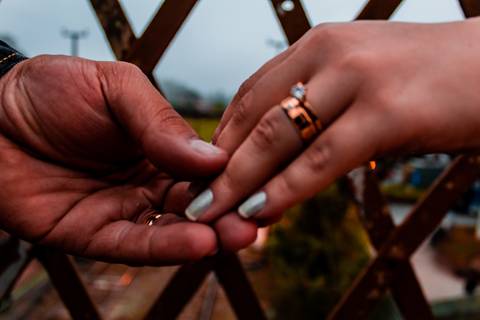 Foto das mãos de um casal, focado nas alianças. Estão em cima de uma passarela, com a linha do trem atrás. '