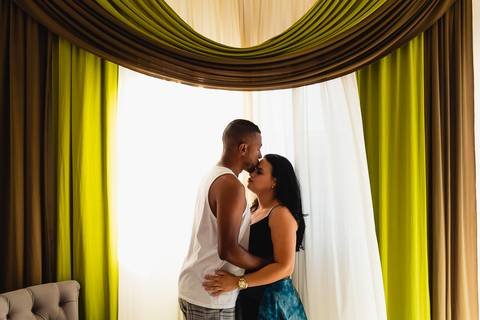 casal em pé,  um de frente para o outro, homem beijando a testa da mulher, na frente de uma cortina verde e branca
'