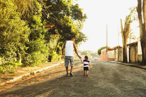 pai e filho caminhando de mãos dadas na rua, é fim de tarde, está um por do sol e tem uma parede com plantas do lado esquerdo '