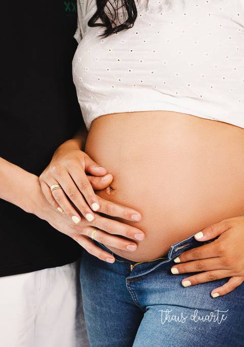 Fotografia de barriga de gestante e mãos com alianças de ouro fotografia por Thais Duarte'