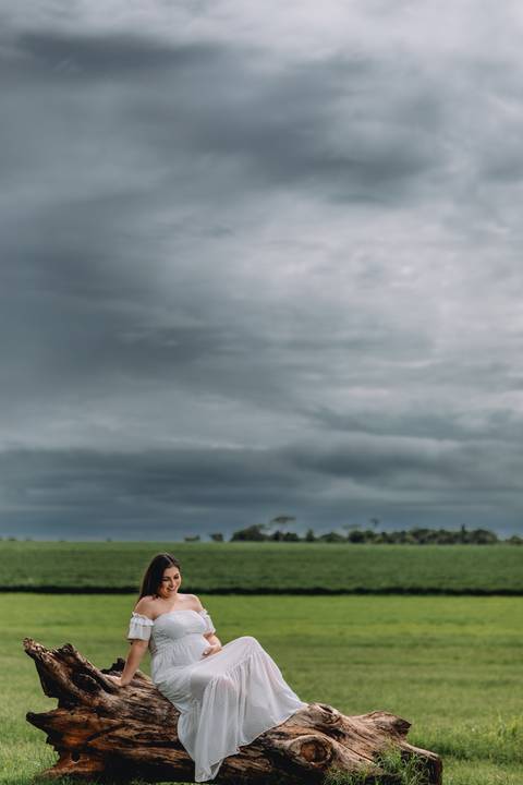 gestante thais sentada em tronco com nuvens carregadas ao horizonte'