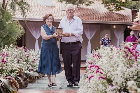 avós da noiva entrando com as alianças e sorrindo noiva emocionada decoração linda casamento ar livre por do sol'