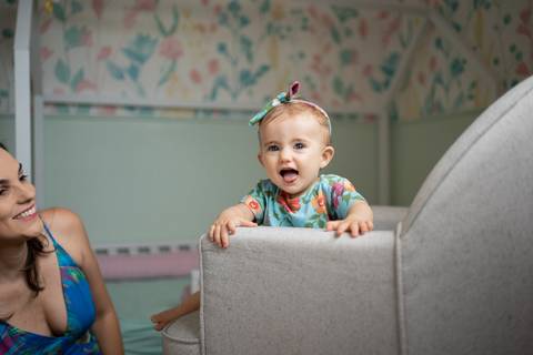 Bebê de 9 meses com lacinho na cabeça, sorrindo para a câmera, sentada na poltrona enquanto a mãe a observa sorrindo; Joao-Francisco-Fotografia; Familia; Niteroi; Rio de Janeiro; Acompanhamento; life-style;'