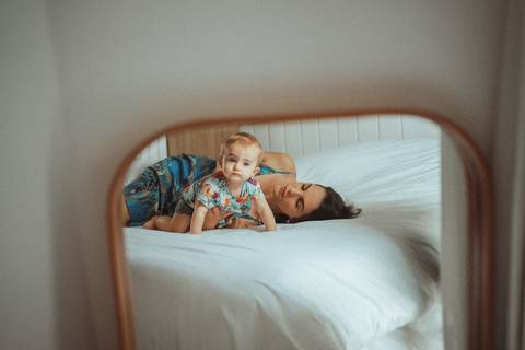Mãe com bebê deitadas sobre edredom branco na cama, refletidas pelo espelho encostado no canto da parede; Joao-Francisco-Fotografia; Familia; Niteroi; Rio de Janeiro; Acompanhamento; life-style;'