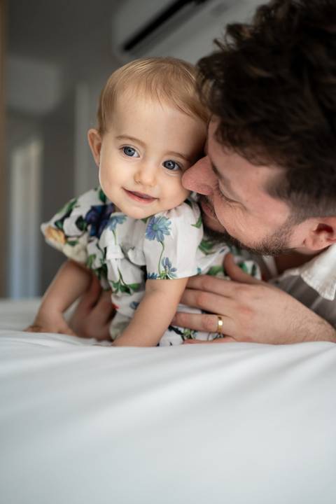 Bebê sentada na cama com leve sorriso no rosto enquanto o pai sorri e cheira bochecha da bebê; Joao-Francisco-Fotografia; Familia; Niteroi; Rio de Janeiro; Acompanhamento; life-style;'