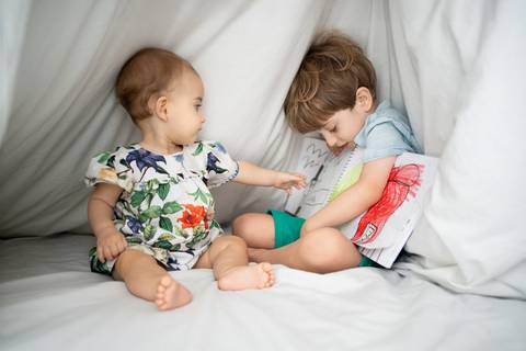 Irmãos dentro de uma cabana feita com edredom; irmão mais velho mostrando o livro de desenhos pra irmã bebê; Joao-Francisco-Fotografia; Familia; Niteroi; Rio de Janeiro; Acompanhamento; life-style;'