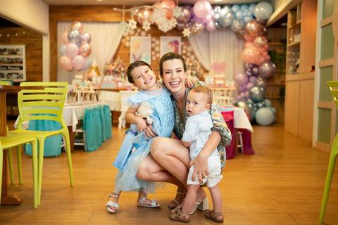 Festa infantil de aniversário de menina de 4 anos com o tema Frozen; princesa Elsa; princesa Anna; vestido azul; mãe agachada abraçada com os filhos, um menino e uma menina; fundo desfocado com decoração balões; quintal de contos; niteroi; Rio de Janeiro;'