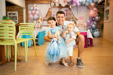 Festa infantil de aniversário de menina de 4 anos com o tema Frozen; princesa Elsa; princesa Anna; vestido azul; pai agachada abraçada com os filhos, um menino; quintal de contos; niteroi; Rio de Janeiro; e uma menina; fundo desfocado com decoração balões'