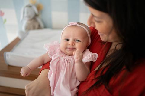 Bebê menina de 3 meses vestindo vestido rosa, olhando para a câmera, no colo da mãe; mãe de roupa vermelha; Joao-Francisco-Fotografia; Familia; Niteroi; Rio de Janeiro; Acompanhamento; life-style;'