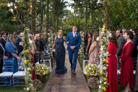entrada do noivo com sua mãe convidados observam de pé decoração natural flores brancas e amarelas'